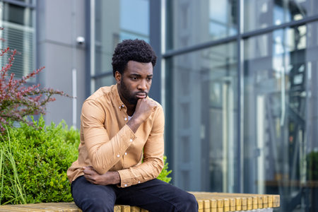 Young man sitting outdoors reflecting on life challenges, experiencing anxiety and feeling sad and worried while contemplating difficult decisions or a stressful situationの写真素材