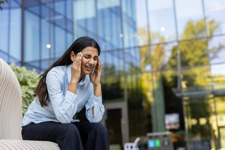 Indian businesswoman sitting outdoors, pressing her temples in pain from a severe headache or...
