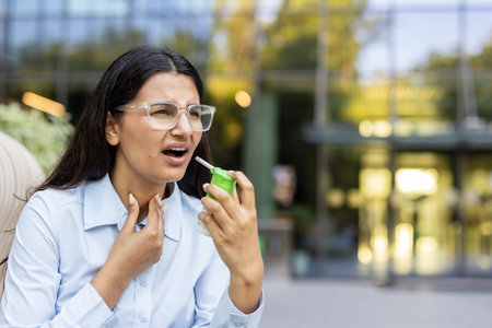 Young woman feeling discomfort from a sore throat, holding her neck and applying throat spray for treatment while standing outdoors with a building in the backgroundの写真素材
