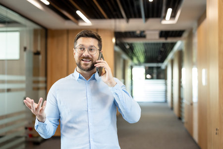 Smiling professional man wearing eyeglasses having a conversation on a mobile phone while standing in a contemporary corporate building hallway, discussing businessの写真素材