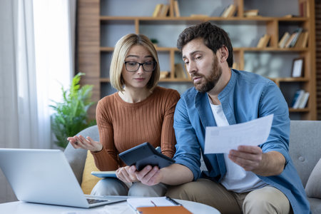 Young couple sitting on a sofa at home, managing household finances, calculating bills, reviewing...