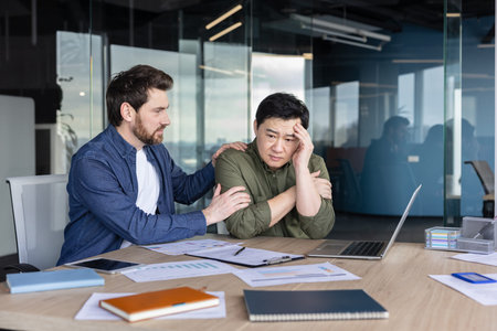 White man comforting a stressed asian coworker in an modern office, offering empathy and counseling...