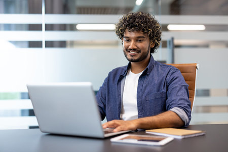 Indian smiling young man sitting in the office at the desk and working, studying on a laptop.