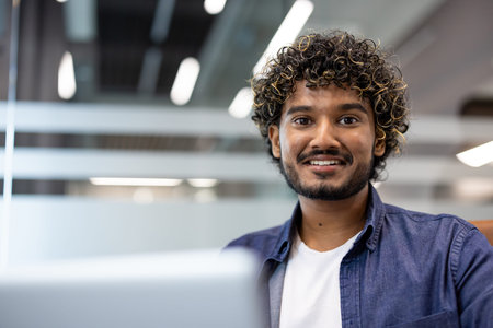 Close-up portrait of a young Indian man working in an office with a laptop and looking at the...