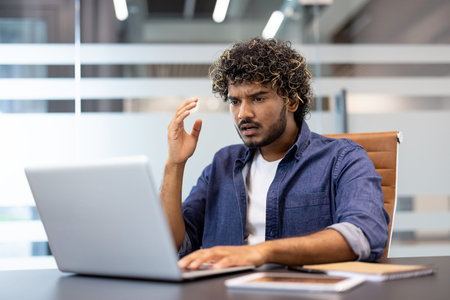 Shocked Indian young man sitting at office desk and looking worriedly at news and problem on laptop.