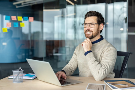 Happy bearded professional man wearing glasses smiling, sitting at desk, using laptop, and looking...