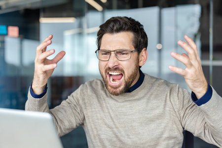 Young man experiencing extreme stress and frustration while yelling at a laptop in a modern office,...