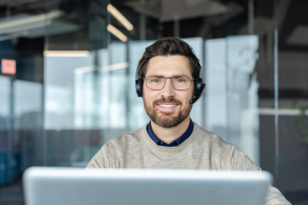 Man wearing glasses and a headset smiling, working at a laptop in a modern office, portraying...