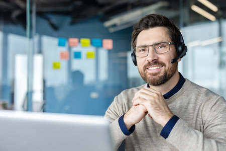 Happy bearded man working in a modern office, wearing a headset with a microphone and glasses,...