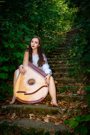 A Ukrainian woman is sitting on the stairs in an old park with a Ukrainian bandura musical...