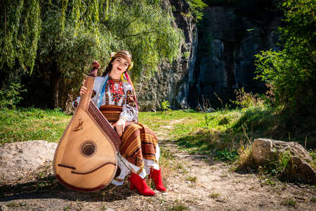 Ukrainian musician in authentic national dress sits by a rock. Ukrainian woman with a bandura musical instrument before performing folk music.の写真素材