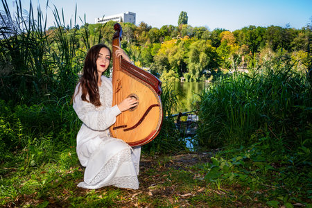 Young Ukrainian woman in national dress plays a bandura by the river on a summer sunny day.