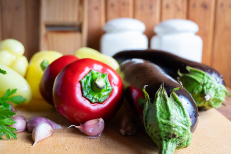 Colorful vegetables, including peppers, eggplants, and garlic, are arranged on a wooden cutting board. The warm atmosphere of the kitchen enhances the freshness of the produce.の写真素材