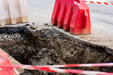 Workers are excavating a deep trench on the road, surrounded by safety barriers. Red and white tape...