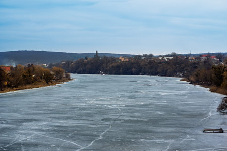 A tranquil river is covered in ice, surrounded by trees and distant buildings. The cloudy sky...