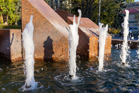 A modern water fountain features several jets shooting water upward in a rhythmic pattern. The sunlight reflects off the water, creating a serene atmosphere in a public park setting.の写真素材