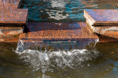Water flows gracefully over a granite block, creating gentle ripples in the tranquil urban fountain. Sunlight reflects off the water, enhancing the peaceful atmosphere of the space.の写真素材