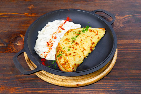 A golden omelette is perfectly cooked and presented on a black skillet, accompanied by a rich, creamy sauce garnished with parsley and spices, set on a wooden tableの写真素材
