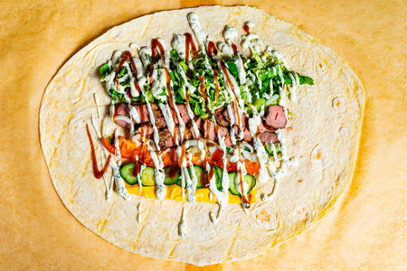 A large tortilla is beautifully arranged with grilled meat, fresh vegetables, and savory sauces. The vibrant colors make it an appetizing meal ready to be rolled and enjoyed.の写真素材