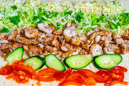 A colorful wrap is being constructed with layers of grilled chicken, lettuce, cucumbers, and red...