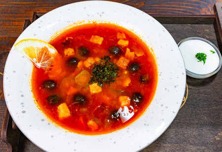 A vibrant bowl of borsch is served, featuring colorful vegetables, garnished with fresh herbs and a lemon slice. A small bowl of cream accompanies itの写真素材