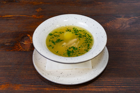 Bowl of clear broth filled with tender chicken pieces, garnished with vibrant green herbs, resting on a rustic wooden table. Perfect for a comforting meal at any time.の写真素材