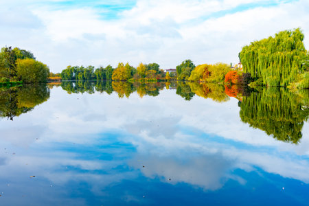 A tranquil lake mirrors vibrant autumn foliage under a blue sky. Lush trees line the shore, creating...