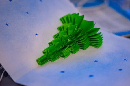 A detailed green paper Christmas tree sits on a white surface dotted with blue snowflakes. The...