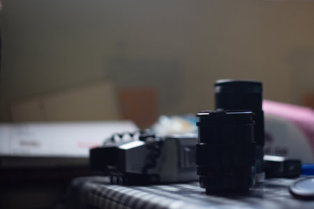 Various camera equipment is scattered on a checkered tablecloth in a softly lit room. The lenses and camera body reflect the ambient light, creating a moody atmosphere.の写真素材