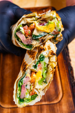 A person wearing black gloves holds a freshly made wrap, showcasing vibrant vegetables, grilled meats, and cheese. The wrap is grilled to a perfect golden brown, resting on a wooden surface.の写真素材