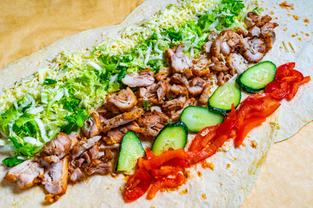 A freshly made wrap is laid out with grilled chicken, shredded cheese, lettuce, cucumbers, and red bell peppers arranged neatly. Vibrant colors and textures create an appetizing display.の写真素材