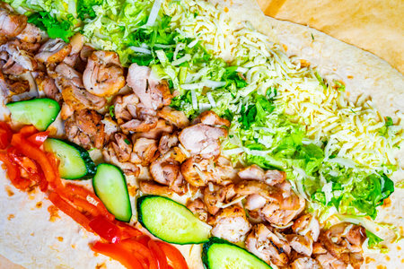 A colorful wrap is prepared with grilled chicken, shredded lettuce, cucumber slices, and cheese,...