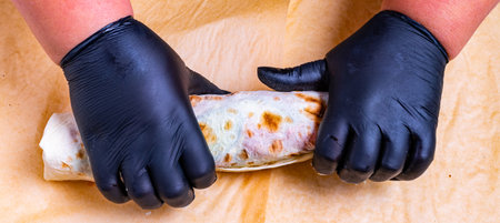 Expert hands in black gloves skillfully roll a freshly cooked tortilla wrap. The texture and color of the wrap indicate a delicious filling within, showcasing culinary craftsmanship.の写真素材