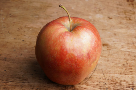 A solitary red apple sits on a rustic wooden counter. Its smooth skin glistens under soft daylight,...
