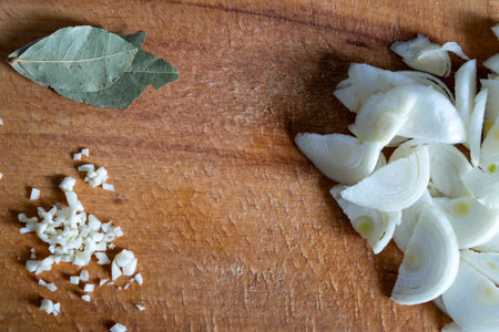 Freshly diced onions are neatly arranged on a wooden cutting board, accompanied by a bay leaf. The ingredients are ready for preparation in a home kitchen setting.の写真素材