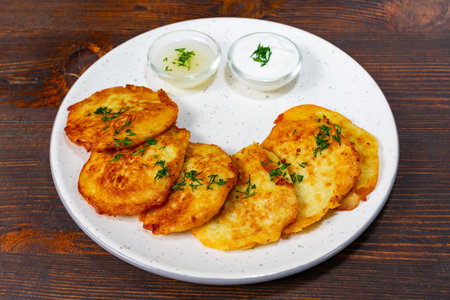 Crispy potato pancakes are plated with two small bowls of creamy sauce and garnished with fresh herbs. The golden-brown treats are perfect for sharing or enjoying alone.の写真素材