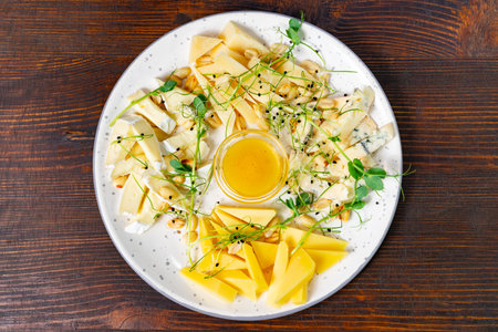 An artistic arrangement of various cheeses alongside honey, nuts, and fresh microgreens is presented on a white plate. The rustic wooden table adds warmth to the dining experience.の写真素材