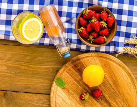 A vibrant display features fresh strawberries, a ripe lemon, and a refreshing drink on a blue and white checkered tablecloth. This picnic setting invites relaxation and enjoyment.の素材