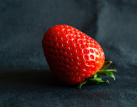 A vibrant red strawberry rests against a dark black fabric background. The natural light emphasizes its glossy surface and detailed seeds, showing its freshness and allure.の素材