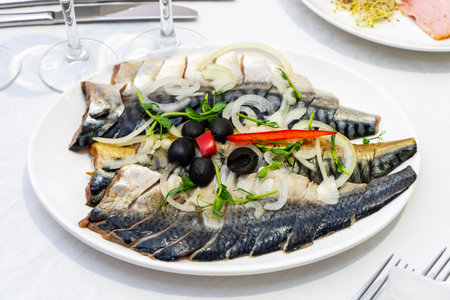 A beautifully arranged plate features fresh mackerel, sliced and topped with onions, olives, and herbs. A red pepper slice adds a pop of color. The setting includes elegant tableware and glasses, suggesting a fine dining experience.の写真素材