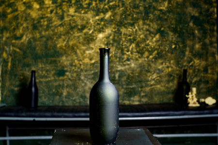 A dark glass bottle stands on a table, surrounded by a textured green and gold wall, creating a...
