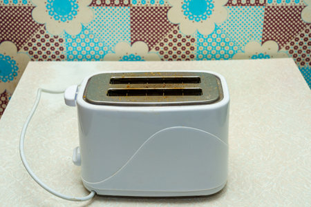 A white two-slice toaster sits on a countertop beside a colorful retro-patterned wall. It is plugged...