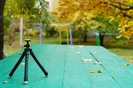 A black tripod stands on a green table covered with yellow leaves. Playground equipment is visible behind it in a park during autumn.の写真素材