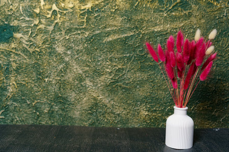 A white vase holds pink flowers on a dark table. Behind it is a textured green wall. This scene shows simple decoration and vivid colors.の写真素材