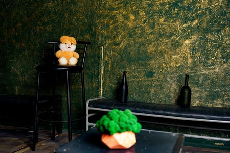 A yellow and brown stuffed animal is on a black stool in a room with dark green walls. There are two black bottles on the bench.の写真素材