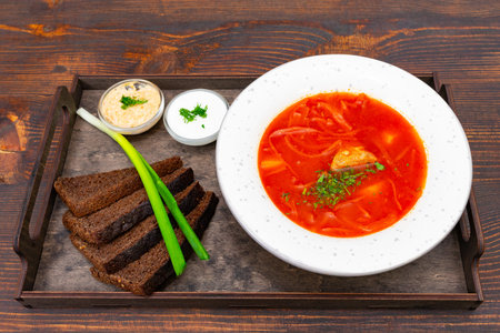 A bowl of Ukrainian red borscht sits on a tray with slices of bread, herbs, and sauces ready for...