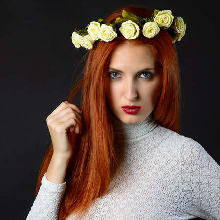 Beautiful young woman with long red hair in wreath of flowers over dark background.の写真素材