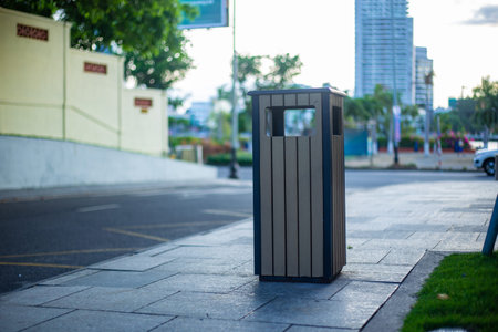 A trash can on the sidewalk next to a bus stop, with a blurred background of the park and building,...