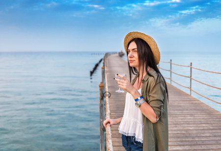 girl in straw hat with glass of wine on the wooden pier on sunset