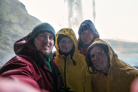 four friends in raincoats make a selfie under an Icelandic waterfall Seljalandsfoss
